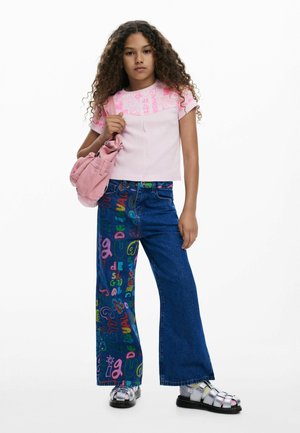 Top de manga corta rosa claro con patrón gráfico, combinado con jeans de pernera ancha con texto colorido. La modelo lleva un bolso rosa y sandalias plateadas.