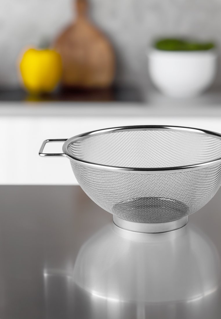 Edelstahl-Mesh-Sieb mit runder Form, zwei Griffen und einem glatten Boden. Im Hintergrund sind eine gelbe Paprika und eine weiße Schüssel zu sehen.