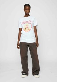 T-shirt en coton blanc avec un imprimé d'une femme blonde dans un cœur rose avec le texte "Blondie" au-dessus. Associé à un pantalon marron ample.