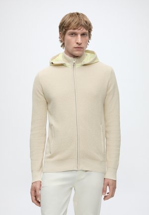 Homme aux cheveux châtain clair portant un pull à capuche beige zippé et un pantalon blanc, debout sur un fond clair uni.