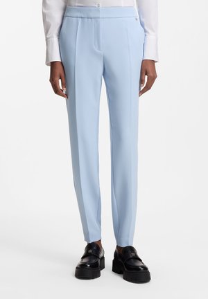 Gerade geschnittene hellblaue Anzughose, kombiniert mit weißem Hemd und schwarzen klobigen Loafers, getragen von einem Model vor weißem Hintergrund.