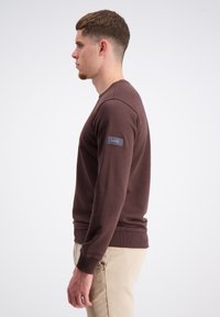 Bruine katoenen sweatshirt met een ronde hals en geribde manchetten; heeft een patch op de linkermouw met logo-details.