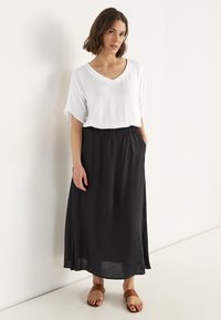 Zwarte lange rok met een elastische tailleband, gecombineerd met een losse witte v-hals top. Bruine slidesandalen maken de outfit compleet.