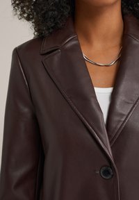 Blazer de cuero burdeos con solapas de muesca, cierre de un solo botón negro y una textura suave; superpuesto sobre una blusa blanca.