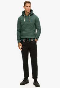 Sudadera verde con capucha y logo texturizado, superpuesta a una camisa de cuadros. Combinada con pantalones negros y botas de cordones negras.