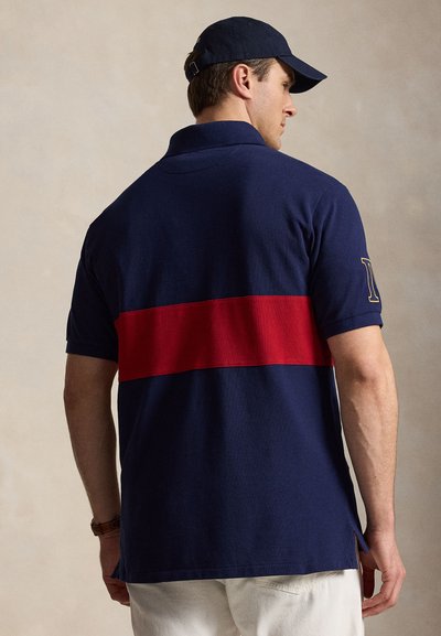 Άνδρας που φοράει μπλε πόλο μπλούζα με ευρεία οριζόντια κόκκινη ρίγα στην πλάτη και ταιριαστό μπλε καπέλο, γυρισμένος πλευρικά μπροστά σε απλό φόντο.