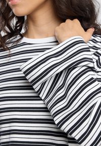 Vrouw met een zwart-wit gestreepte lange mouwen top, met de hand omhoog bij de schouder en krullend haar zichtbaar.