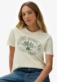 Kremowa koszulka z bawełny z krótkimi rękawami, z okrągłym zielonym wzorem graficznym, który zawiera tekst oraz motyw misia.