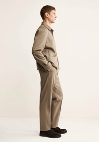Giacca marrone e pantaloni abbinati con vestibilità su misura, realizzati in tessuto liscio. Il modello si trova di lato, con le mani nelle tasche, indossando scarpe scure.