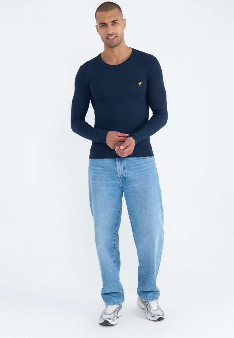 Mørkeblå langermet t-skjorte i kombinasjon med lyseblå jeans. Har en tettsittende design, glatt tekstur og en liten gul logo på brystet.