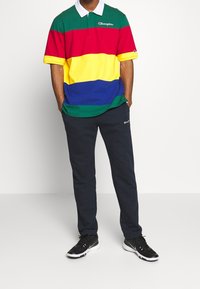 Polo coloré avec des rayures horizontales vertes, rouges, jaunes et bleues, avec un col blanc. Assorti à un pantalon marine foncé et des chaussures noires.