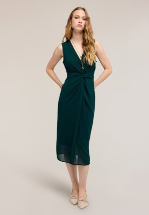 Kvinde står med hænderne på hofterne iført en ærmeløs mørkegrøn midi-kjole med en indsnævret talje og beige spidse sko.