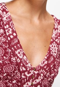 Rotes Kleid mit Muster, das eine tiefe V-Ausschnitt, Knopfdetails und florale Designs in kontrastreichem Weiß aufweist. Weiche Stofftextur sichtbar.