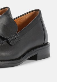 Μαύρο δερμάτινο μοκασίνι με στρογγυλεμένη μύτη και διακοσμητικό φιόγκο. Ο εσωτερικός χώρος είναι καφέ, με χαμηλό τακούνι τύπου block και λεία υφή.