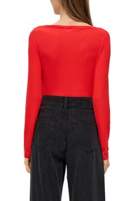 Rotes Langarmshirt mit Rundhalsausschnitt, kombiniert mit hoch taillierten schwarzen Jeans mit zwei Gesäßtaschen und einem sichtbaren Taillenlogo.