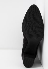 Musta mokkanahkainen nilkkaboots, joka lepää kyljellään, näyttäen terävän pohjan, jossa on geometrinen urituskuvio ja matala lohkare-korko.