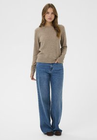 Beżowy sweter wełniany z okrągłym dekoltem i ściągaczem, w połączeniu z dżinsami w stylu wide-leg z wysokim stanem w średnim niebieskim kolorze. Brązowe buty.