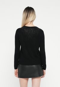 Un pull noir à manches longues, avec un décolleté arrondi et un motif pailleté au dos. Assorti à une mini-jupe en cuir noir.