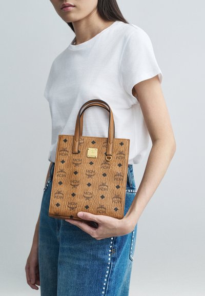 Nainen, jolla on päällään valkoinen t-paita ja niittikuvioiset siniset farkut, pitää tan-väristä MCM-laukkua, jossa on musta logo-kuvio ja kultainen plaque, rinnan korkeudella.