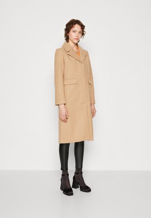Beige ullblandingsfrakk med notch-krage, lange ermer og to frontlommer, båret over svarte fake skinnleggings og heeled ankelstøvler.