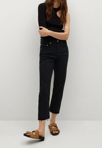 Jeans negros de cintura alta con dobladillo deshilachado, combinados con sandalias de tiras marrones. La parte superior es un suéter negro ajustado con un corte asimétrico.