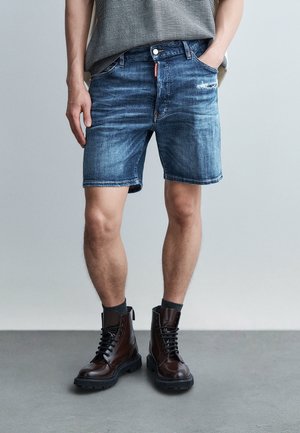 Moški, oblečen v izbljuvane modre denim kratke hlače, črne nogavice in temno rjave čevlje z vezalkami, stoji z eno roko v žepu.