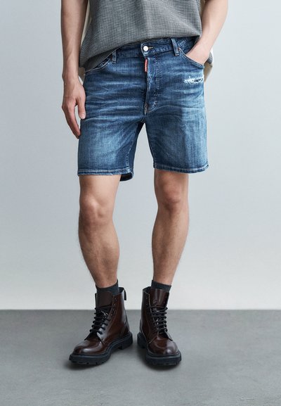 Homme portant un short en jean bleu délavé, des chaussettes noires et des bottes marron foncé à lacets, debout avec une main dans la poche.