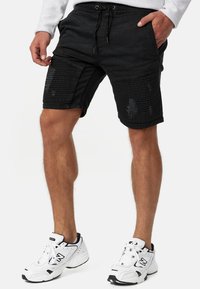 Mężczyzna ubrany w czarne prążkowane spodenki rowerowe z przetarciami, białe sneakersy i jasnoszary długi rękaw, pozujący z rękami blisko kieszeni.