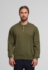 Man with beard wearing olive green long-sleeve polo shirt and navy pants, standing against a plain light gray background.