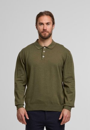 Uomo con barba che indossa una polo a maniche lunghe verde oliva e pantaloni blu navy, in piedi davanti a uno sfondo grigio chiaro uniforme.