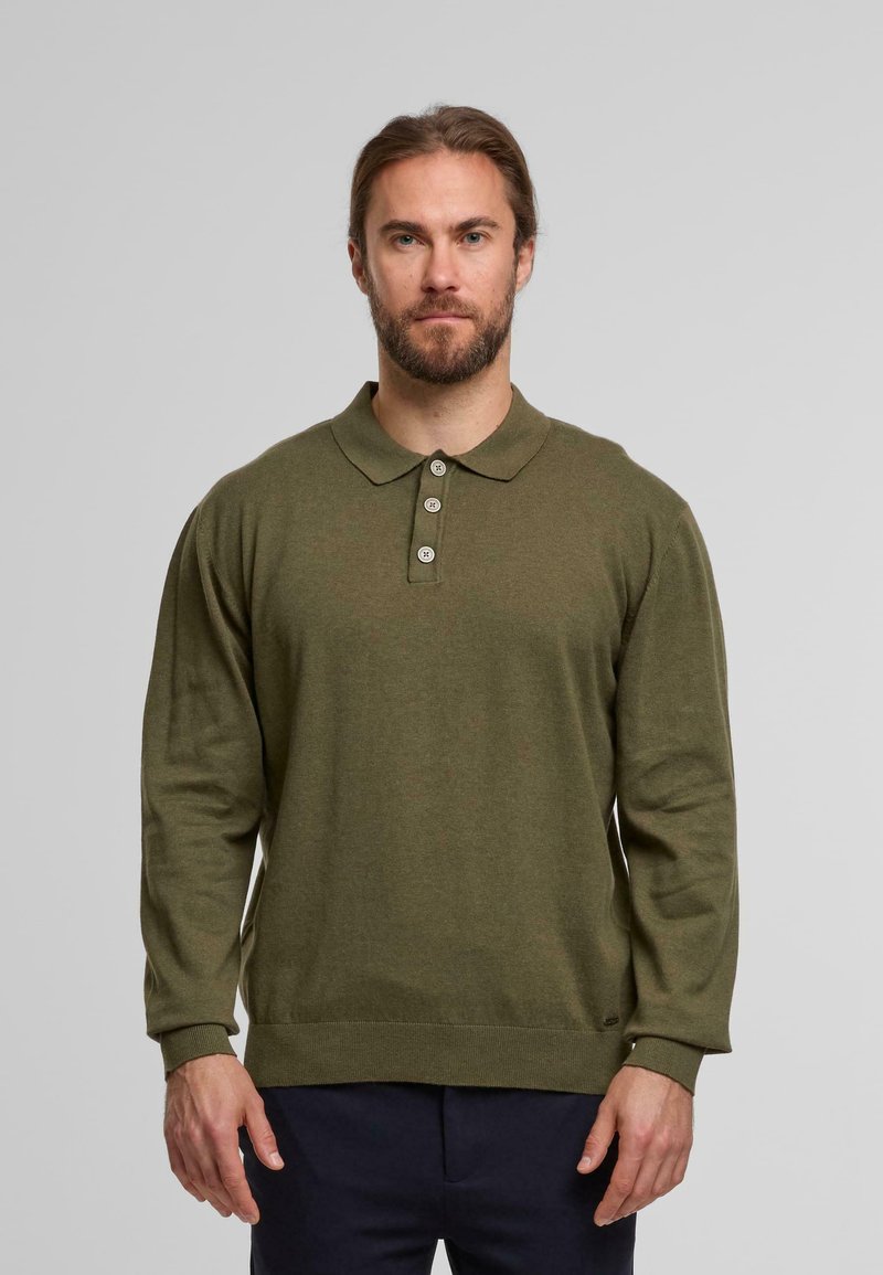Uomo con barba che indossa una polo a maniche lunghe verde oliva e pantaloni blu navy, in piedi davanti a uno sfondo grigio chiaro uniforme.