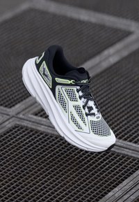White and black running shoe with mesh upper, neon green accents, thick sole, and black laces. Features a padded collar and breathable design.