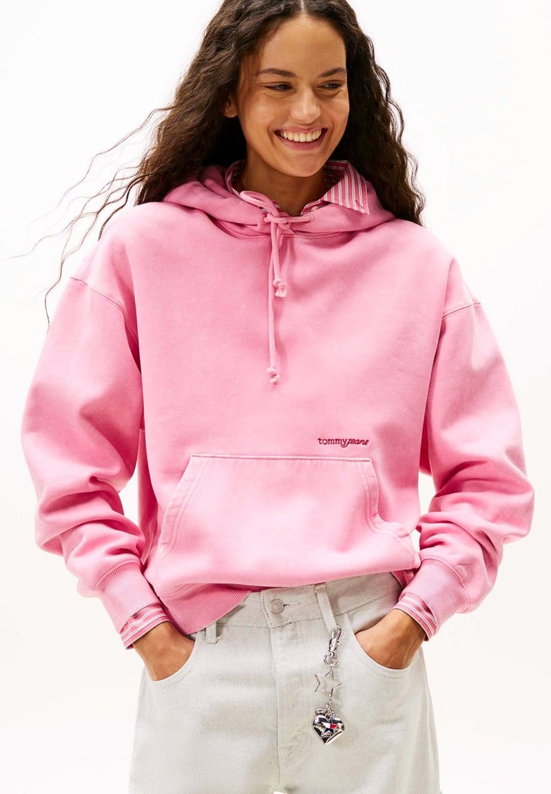 Felpa rosa con cappuccio, tasca frontale e coulisse. Presenta un collo interno a righe e un ricamo con logo. Abbinata a jeans chiari.