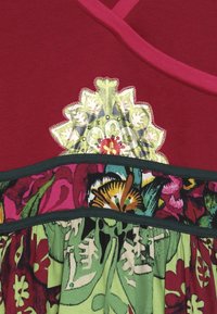 Czerwony top z głębokim dekoltem w serek i haftem z cekinami w kwiaty. Pod spodem zielony materiał w wzory z różowymi i zielonymi kwiatami. Czarna lamówka jako akcent.