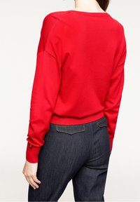 Röd stickad tröja med avslappnad passform och ribbade kanter; bärs med mörkblå denimbyxor. Tröjan har subtila stygninjer.