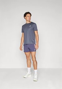 Conjunto atlético masculino composto por uma camisa azul-marinho de mangas curtas e calções roxos, feitos de tecido leve, com um detalhe de logótipo no peito.