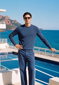 Homme portant un pull bleu et un pantalon avec des lunettes de soleil, debout près de la rambarde près de la piscine et de l'océan avec des montagnes en arrière-plan.