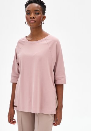 Zwarte vrouw met kort haar, gekleed in een lichtroze losvallende top met driekwart mouwen en beige broek, staand tegen een witte achtergrond.