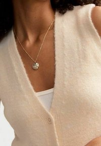 Kremowy, bezrękawnik z dekoltem w serek, miękki w dotyku, z dwoma guzikami, noszony na białym topie na ramiączkach, zestawiony z naszyjnikiem z złotą zawieszką.