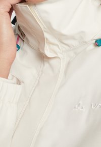 Veste légère beige avec col montant, tissu texturé, poignets élastiques et cordons de serrage colorés aux accents bleus et roses.