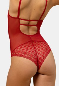 Un body rouge en tissu en mesh transparent, avec des bretelles de soutien, un dos dégagé et un ourlet en dentelle complexe avec un motif géométrique.