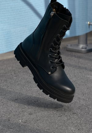 Bottine de combat en cuir noir avec semelle épaisse et fermeture éclair latérale, flottant au-dessus d'un sol en bitume avec un fond bleu.