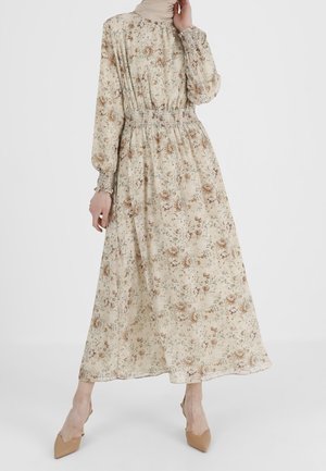Kvinde iført en lang, beige blomsterkjole med smock i taljen og manchetter, kombineret med brune spidse hæle, poserer foran en ensfarvet baggrund.