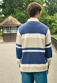 Long-sleeve polo shirt in navy blue and cream stripes, featuring a collared neckline and a relaxed fit, worn with light blue jeans.