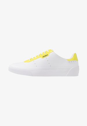 Zapatilla blanca con un acento amarillo brillante, diseño de cordones y suela de goma texturizada. Logo de la marca exhibido en el lateral.