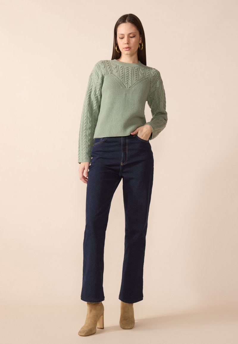 Maglione verde a maglia rasata con texture a coste, abbinato a jeans dritti blu scuro e stivali alla caviglia beige.