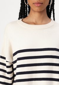 Cream-colored knit sweater with navy horizontal stripes. Features a ribbed neckline and loose fit, showcasing soft texture.