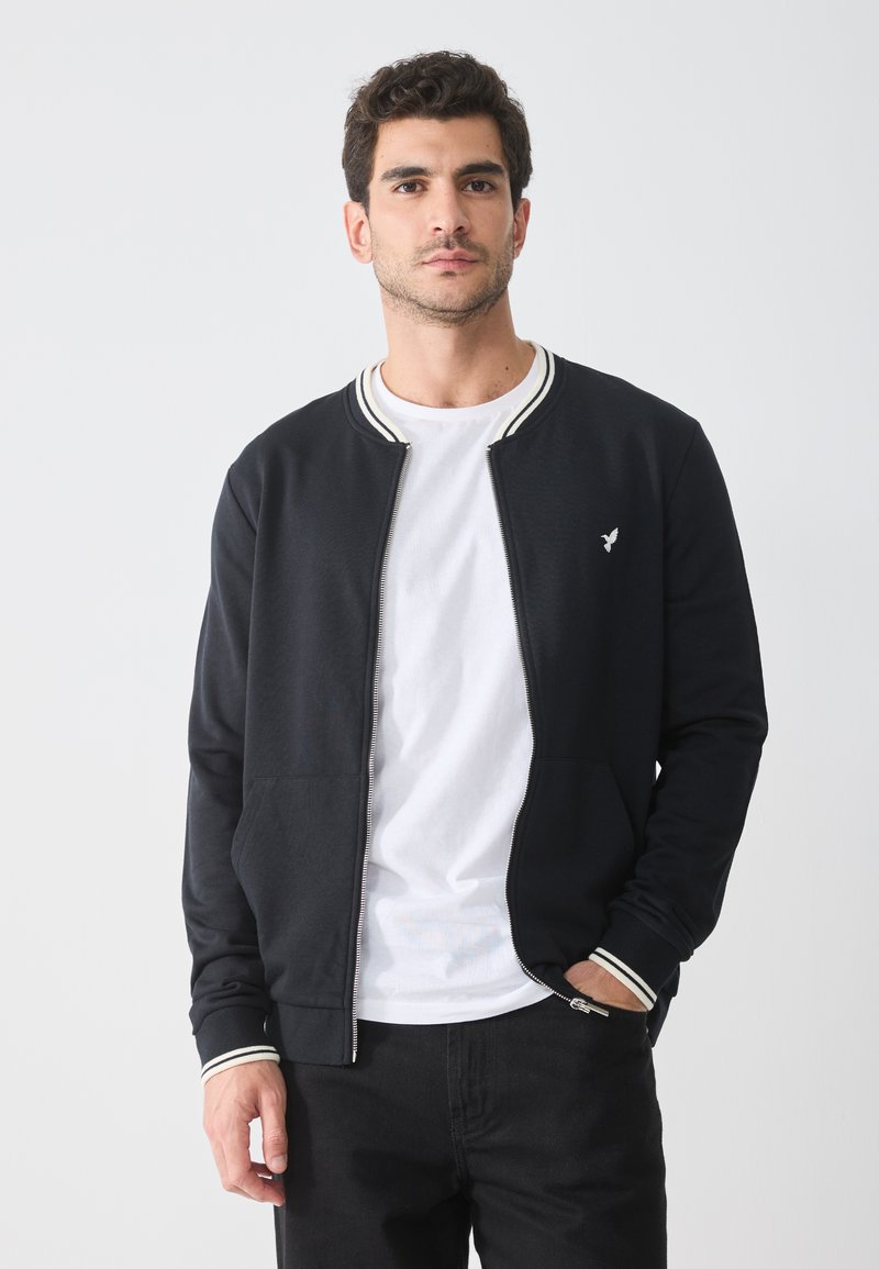 Black zip-up jacket with white striped collar and cuffs, front pockets, and an embroidered logo. Worn over a white T-shirt and black pants.
