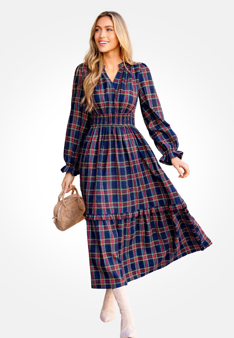 Vestido maxi de cuadros marino con mangas largas, cintura fruncida, dobladillo con volantes y cuadros multicolores. La modelo sostiene un bolso tan.