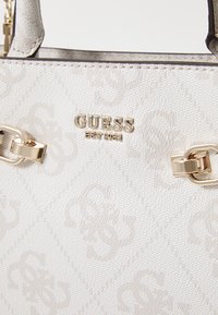 Borsa bianca testurizzata con hardware dorato, caratterizzata dal logo "GUESS" e da un delicato motivo a diamante. Cinghie attaccate con fermagli decorativi.